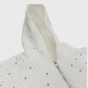 May include: White hooded sweatshirt with a silver zipper and scattered rhinestones. The hoodie features a soft, white interior and a full-length zipper. The rhinestones are evenly spaced across the fabric.