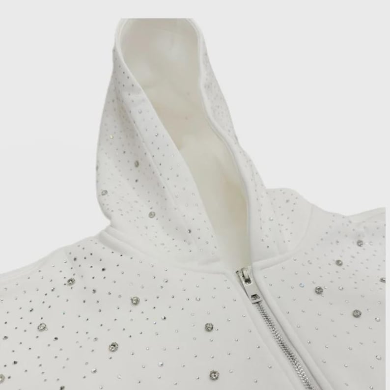 May include: White hooded sweatshirt with a silver zipper and scattered rhinestones. The hoodie features a soft, white interior and a full-length zipper. The rhinestones are evenly spaced across the fabric.