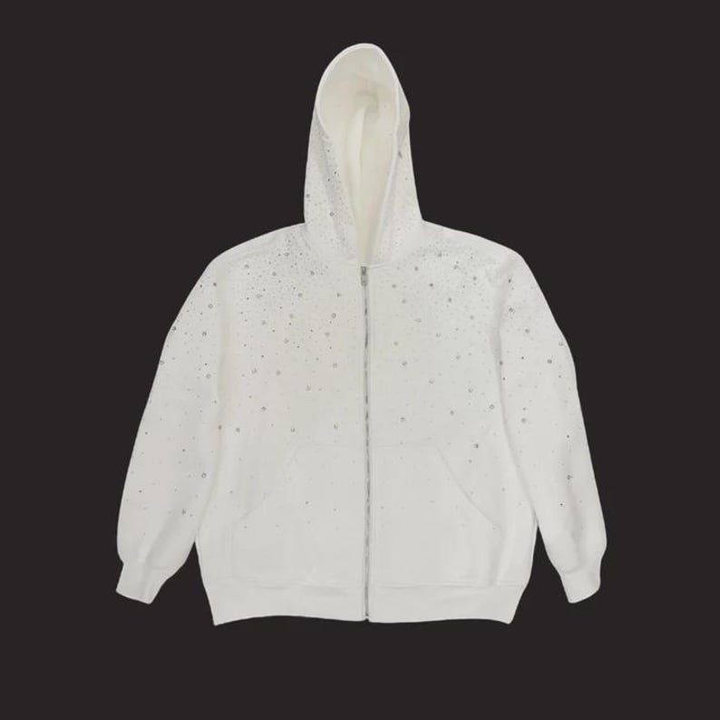 May include: White zip-up hoodie with a drawstring hood and front pockets. The hoodie is embellished with scattered silver-colored rhinestones. The zipper is silver-colored. The hoodie is made of a soft material.