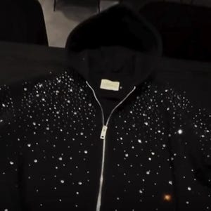 May include: Black hooded sweatshirt with a full-length zipper and a scattering of sparkling embellishments. The sweatshirt has a drawstring hood and a small tag near the neckline. The design is a dark, elegant style.