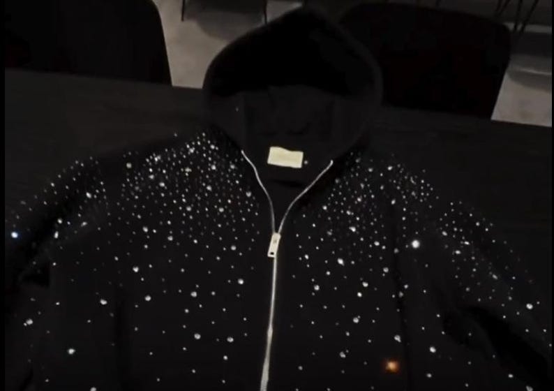 May include: Black hooded sweatshirt with a full-length zipper and a scattering of sparkling embellishments. The sweatshirt has a drawstring hood and a small tag near the neckline. The design is a dark, elegant style.