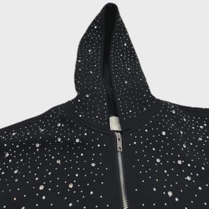 May include: Black zip-up hoodie adorned with numerous sparkling rhinestones. The hood and front of the jacket are densely covered in the embellishments. The zipper is silver. The hoodie is a casual garment.