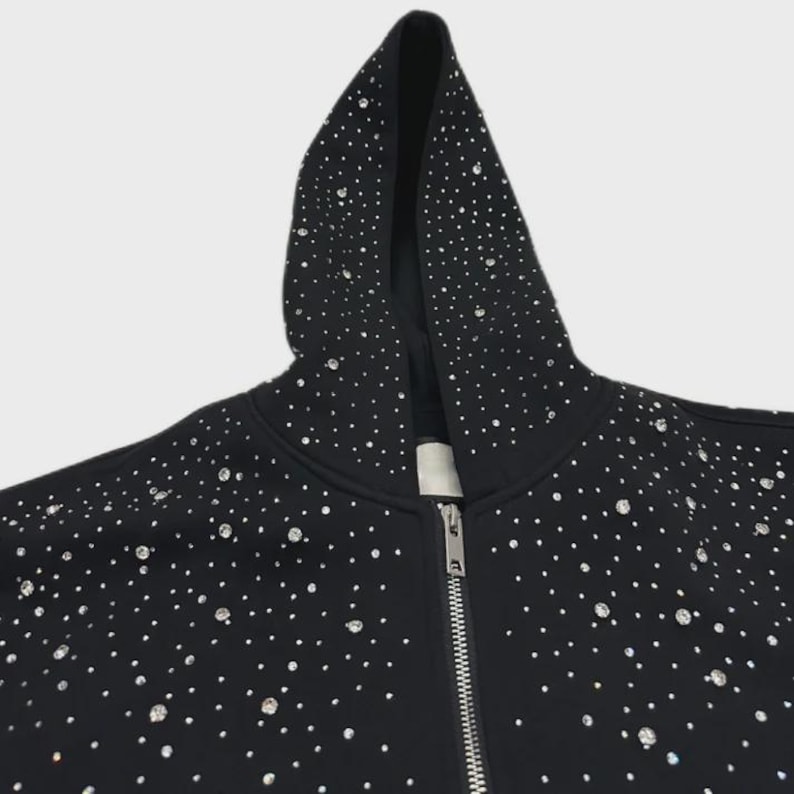 May include: Black zip-up hoodie adorned with numerous sparkling rhinestones. The hood and front of the jacket are densely covered in the embellishments. The zipper is silver. The hoodie is a casual garment.