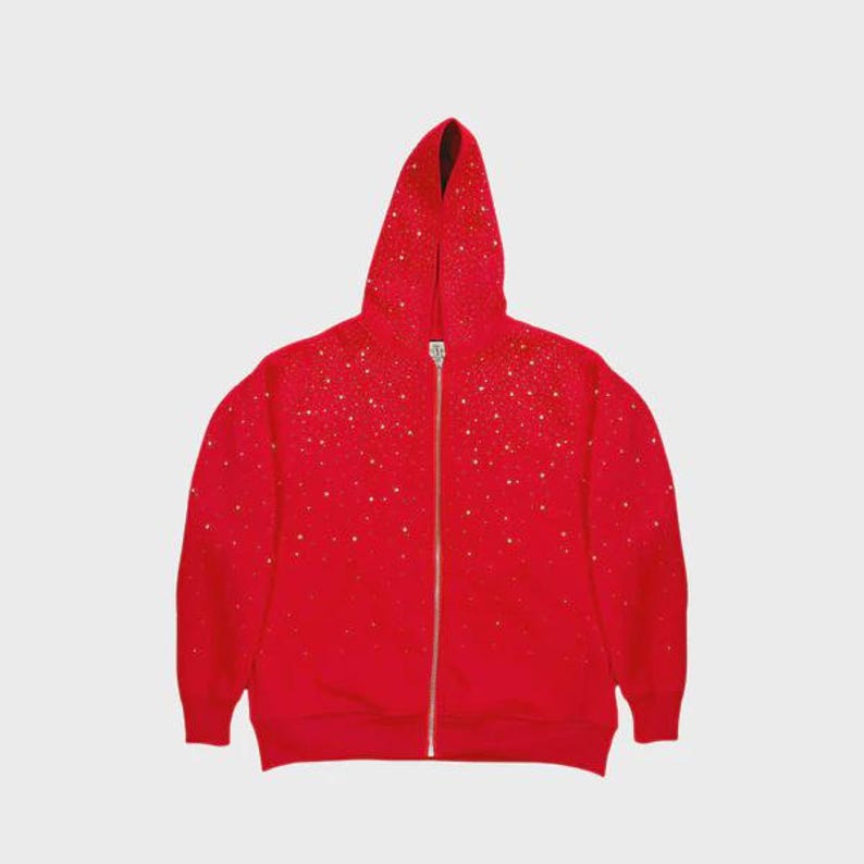 May include: A red hooded zip-up sweatshirt with a scattering of gold-colored embellishments. The sweatshirt has a full-length zipper and a hood. The fabric appears to be a soft material. The background is a solid, light gray color.
