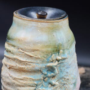 May include: A ceramic jar with a lid, featuring a textured, uneven surface with a glaze transition from light blue and green to a sandy beige. The base is dark brown, and the lid is a smooth, dark charcoal gray with a small central knob.