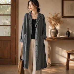 May include: A long, charcoal gray button-down coat with a notched lapel, long sleeves, and two large front patch pockets. The coat is worn over a black tank top and light beige wide-leg trousers, paired with a brown tote bag held at the side.