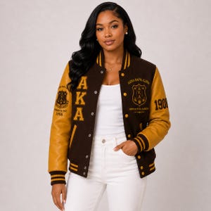 May include: A brown and gold letterman jacket with the letters "AKA" and "1908" embroidered on it. The jacket has gold leather sleeves and a button-up front. The jacket also has a crest on the chest.
