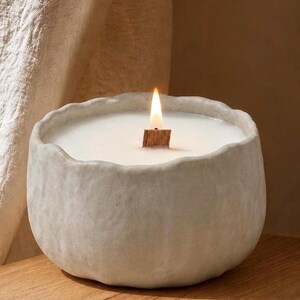 Peut inclure: Une bougie blanche dans un bol en céramique gris clair, rond et texturé. La bougie possède une mèche en bois unique qui est allumée, produisant une petite flamme vive. L'extérieur du récipient en céramique présente un bord irrégulier.