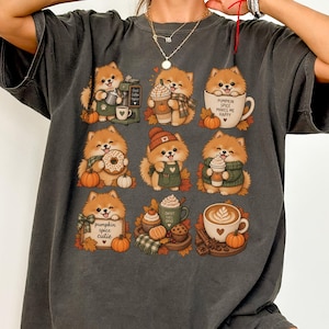 May include: A charcoal gray oversized t-shirt featuring a graphic print of nine illustrations of Pomeranian dogs with coffee, pumpkins, and autumn themes. One illustration includes the text "PUMPKIN SPICE MAKES ME HAPPY" and another says "pumpkin spice cutie".