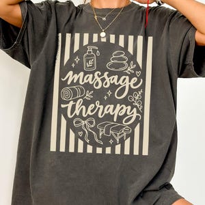 May include: A charcoal gray short-sleeve t-shirt featuring a cream-colored graphic with vertical stripes and a central circle containing the text massage therapy, surrounded by illustrations of a lotion bottle, stones, a towel, and a table.