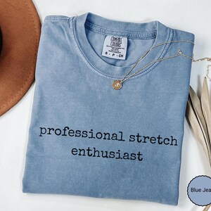 May include: A blue jean colored t-shirt with a crew neckline and short sleeves. The shirt features the black text "professional stretch enthusiast" printed on the front. A gold-toned necklace with a small circular pendant rests on the collar.