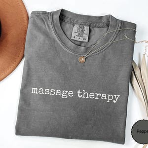 May include: A charcoal gray short-sleeve t-shirt with the text massage therapy printed in white across the chest. The shirt features a Comfort Colors brand label at the inner neckline with size S-P-CH and is made of 100% cotton fabric.