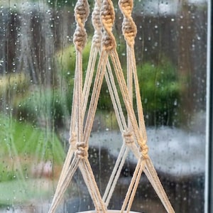 May include: A macrame plant hanger with a white ceramic bowl. The beige rope is knotted in a spiral design at the top and forms a triangular support for the bowl. The bowl is suspended in front of a window with raindrops.