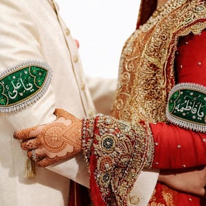 May include: Two armbands featuring green fabric with white Arabic calligraphy and intricate beaded borders. One armband displays the text Ya Ali and the other displays Ya Fatima. These decorative pieces are worn on the sleeves of formal wedding attire.