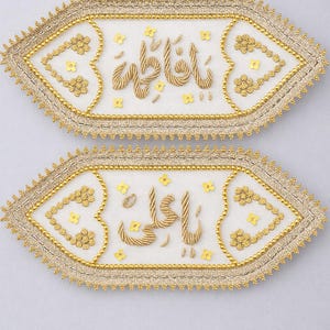 Könnte beinhalten: Zwei dekorative, weiße Stoffstücke mit goldener Stickerei und dem arabischen Schriftzug "Fatima". Jedes Stück hat einen Perlenrand, florale Akzente und einen gewellten Rand.