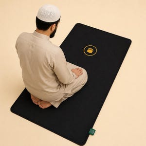 Op de afbeelding: Een rechthoekig zwart gebedskleed met een centraal geplaatst goudkleurig cirkelvormig embleem dat de Kaaba afbeeldt. Het kleed heeft afgeronde hoeken en een klein groen rechthoekig label aan de rechteronderkant voor een strakke uitstraling.
