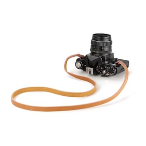May include: A black 35mm film camera with a lens attached, featuring a tan leather neck strap with visible stitching. The camera body includes various silver dials, a shutter button, and a hot shoe mount on the top surface.