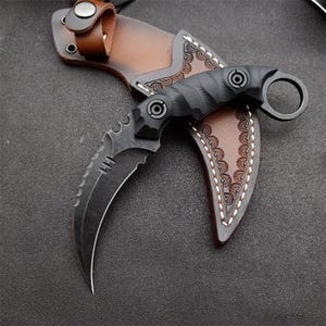 Puede incluir: Un cuchillo karambit negro con una hoja curva y un anillo en el extremo del mango. El cuchillo descansa sobre una funda de cuero marrón con costuras decorativas. El mango tiene un agarre texturizado.