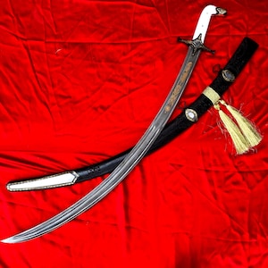 Puede incluir: Una espada curva con empuñadura blanca y detalles dorados en la hoja. La espada descansa sobre una tela roja, con una vaina negra con borlas doradas y detalles decorativos.