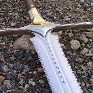 May include: A detailed image of a sword with a silver blade and a brown handle. The blade has a serrated edge and an inscription. The hilt is gold-coloured. The sword is lying on a surface of small rocks.