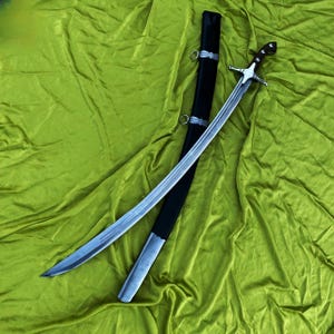 May include: A curved sword with a silver blade and a wooden handle, resting on a green cloth. The sword is accompanied by a black sheath with silver accents. The blade is approximately 76 cm long.