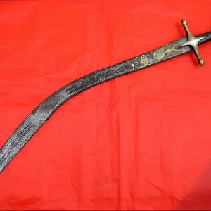 Puede incluir: Un sable curvo con una hoja oscura y texturizada y una empuñadura negra. La empuñadura presenta una guarda y detalles dorados. La hoja tiene grabados decorativos sobre un fondo rojo.