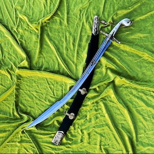 May include: A curved sword with a light blue blade and a white handle, resting on a black sheath with gold-coloured accents. The sword is displayed against a green, textured background.