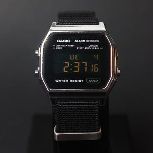 Peut inclure: Une montre numérique Casio avec un cadran noir et un bracelet en tissu noir. La montre affiche l'heure, le jour et la date. Le cadran de la montre porte l'inscription "CASIO ALARM CHRONO" et "WATER RESIST WR".