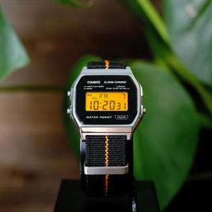 May include: A digital wristwatch with a silver-tone rectangular case and a black nylon strap featuring a central orange stitched line. The watch face displays a bright yellow digital screen showing the time 10:20, the date, and text including CASIO, ALARM CHRONO, and WATER RESIST.