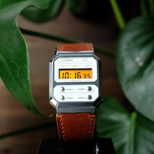 May include: A Casio digital watch with a black rectangular case and a brown leather strap. The watch face features an orange digital display showing 10:16:35, with the text CASIO, WR, and ILLUMINATOR printed on the white and black frame.