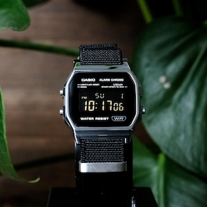 May include: A digital Casio wristwatch with a silver-tone rectangular case and a black nylon strap. The black display shows the time 10:17:06, the day SU, and the date 1. Text on the face includes CASIO, ALARM CHRONO, Lithium, WATER RESIST, and WR.
