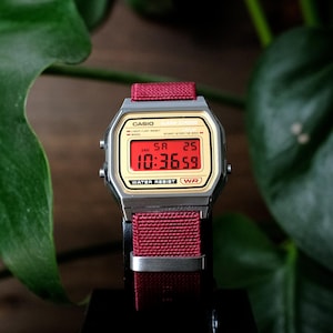 May include: A digital wristwatch with a silver-tone rectangular case and a gold-tone face. The display shows red digital numbers reading 10:36:59, the day SA, and the date 25. It features a dark red woven fabric strap with a silver metal buckle.