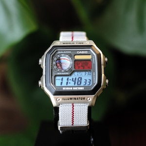 May include: A Casio digital wristwatch with a silver-tone rectangular case and a light gray fabric strap featuring a thin red vertical stripe. The watch face displays WORLD TIME, 10 YEAR BATTERY, and ILLUMINATOR text with a digital time readout.