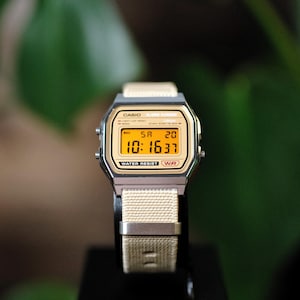 Peut inclure: Une montre-bracelet numérique avec un boîtier rectangulaire argenté et un bracelet en tissu tissé beige. Le cadran présente un affichage numérique teinté de jaune indiquant l'heure 10:16:37, la date SA 20 et le texte CASIO ALARM CHRONO WATER RESIST.