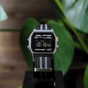 May include: A digital Casio Alarm Chrono watch with a silver-tone rectangular case and a black digital display. The watch features a black fabric strap with two vertical grey stripes and silver-tone metal hardware. The screen displays 10:16.
