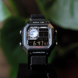 May include: A black Casio digital watch with a square face, featuring a world time display, a moon phase indicator, and a 10-year battery. The watch has a black nylon strap, four side buttons, and the text Illuminator printed on the bottom bezel.