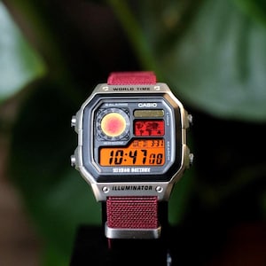 May include: A Casio digital watch with a silver-tone rectangular case and a maroon fabric strap. The watch face features a world time map, a circular analog-style display, and an orange digital screen showing the time 10:47 and date 3-31.