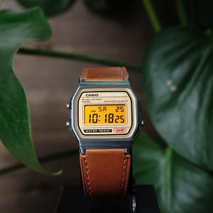 May include: A digital wristwatch with a metallic gunmetal gray case and a tan leather strap. The watch face features a gold-toned background with a digital display showing the time 10:18, the date 25, and the day SA. Text includes CASIO, ALARM CHRONO, and WATER RESIST.