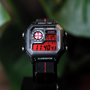 May include: A black digital wristwatch with a red display, featuring the text WORLD TIME, CASIO, CORP., and ILLUMINATOR. The watch face includes a circular red and white umbrella graphic, a world map display, and a black fabric strap with a red center stripe.