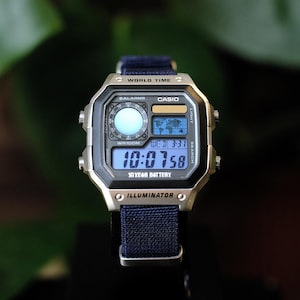 May include: A digital wristwatch with a silver-tone rectangular case and a dark blue fabric strap. The face features a world map display, a circular analog-style sub-dial, and a digital time readout showing 10:07:58. Text includes WORLD TIME, CASIO, 5 ALARMS, WR100M, 10 YEAR BATTERY, and ILLUMINATOR.