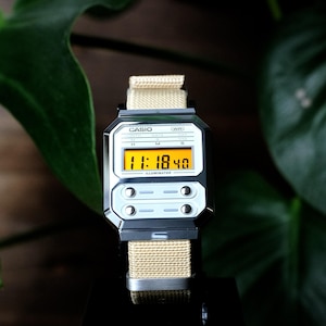 May include: A digital wristwatch with a rectangular silver-tone case and a beige nylon strap. The watch face features a bright orange digital display showing 11:18:40, with the text CASIO, WR, and ILLUMINATOR printed on the white dial surface.