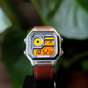 May include: A Casio digital watch with a silver-tone rectangular case and a brown leather strap. The watch face features a world map display, a circular analog-style sub-dial, and a bright orange digital screen showing the time 10:52:02.