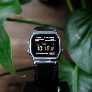 May include: A digital wristwatch with a silver-tone rectangular case and a black leather strap. The black watch face displays the time 10:18, the date, and the text CASIO ALARM CHRONO, LITHIUM, and WATER RESIST WR in white lettering.