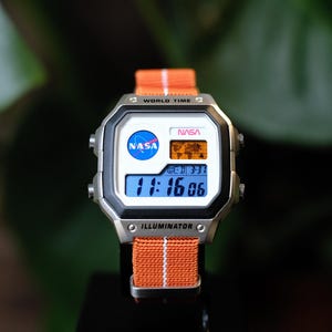 May include: A digital wristwatch with a silver-tone rectangular case and an orange fabric strap featuring a thin white center stripe. The watch face displays the NASA logo, the text WORLD TIME, NASA, and ILLUMINATOR, and a world map graphic.