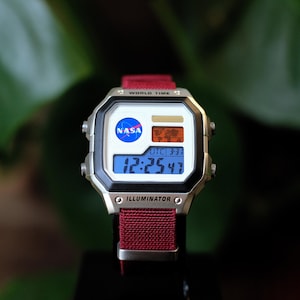 May include: A digital wristwatch with a silver-tone rectangular case and a dark red fabric strap. The watch face features the NASA logo, a world map graphic, and a digital display showing the time 12:25:47, with the text WORLD TIME and ILLUMINATOR.