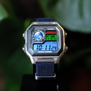 May include: A Casio digital watch with a silver-tone square case and a dark blue fabric strap. The watch face features a world map display, a circular globe graphic, and a digital screen showing the time 10:11:24 and the date SUN 3-31.