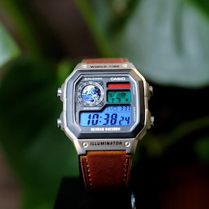 May include: A Casio digital watch with a silver-tone square case and a brown leather strap. The watch face features a world map display, a circular globe graphic, and a digital time readout. Text on the watch includes World Time and Illuminator.