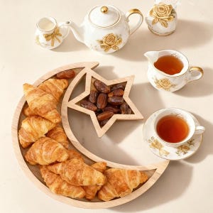 May include: A crescent moon and star shaped wooden serving tray filled with golden brown croissants and dark brown dates, accompanied by a white porcelain tea set featuring gold rose and ribbon patterns, including a teapot, creamer, and teacup.