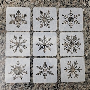 May include: Nine square stencils with different snowflake designs. Each stencil features a unique snowflake pattern in shades of gray and silver. The stencils are arranged in a grid pattern on a speckled, dark surface.