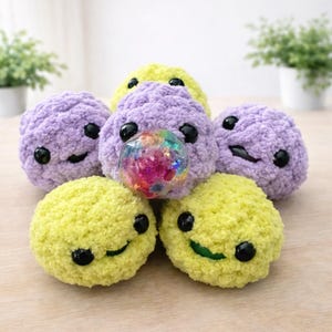 May include: A collection of handmade, plush, smiling creatures in shades of purple and yellow. Each has black button eyes and a stitched mouth. One purple creature has a clear, glitter-filled sphere. The toys are arranged on a light wooden surface.